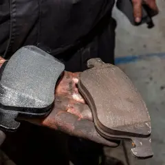 Mani sporche di grasso tengono due pastiglie dei freni: una nuova e una usurata. Un modo per capire come controllare pastiglie freni senza smontare ruota.
